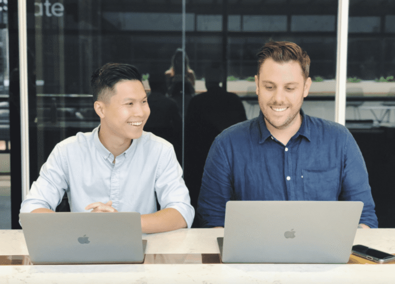 Two men, one Asian and one Caucasian, sitting at a table smiling while working on laptops in a modern accounting firm in Melbourne. | Future Advisory