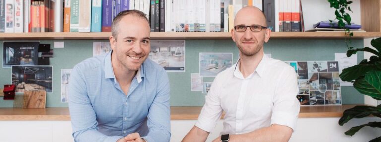 Two men smiling at the camera, seated at a table in an accounting firm in Melbourne with books and plants in the background. One is wearing a blue shirt and the other a white shirt. | Future Advisory